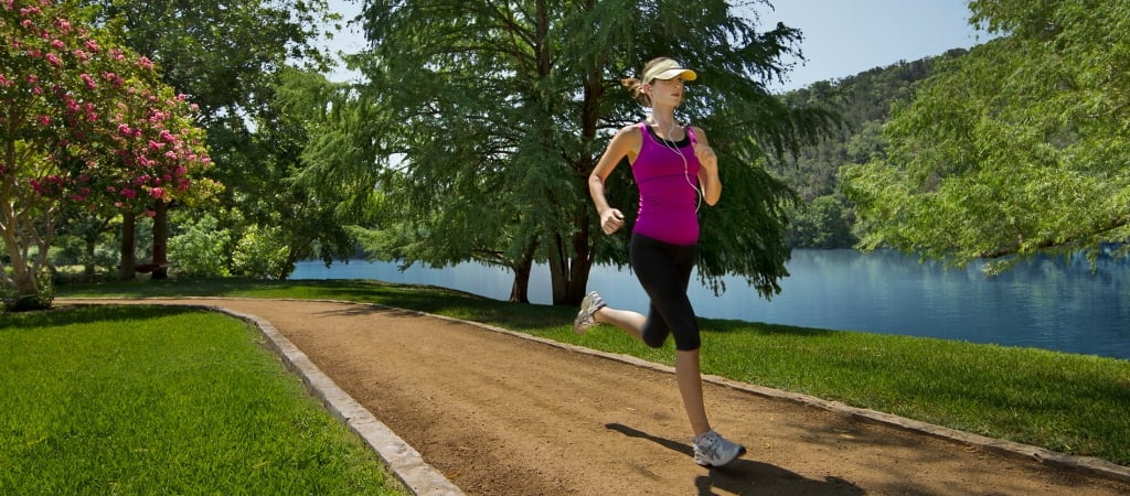 A woman in athletic wear runs along a dirt path surrounded by trees and a lake.