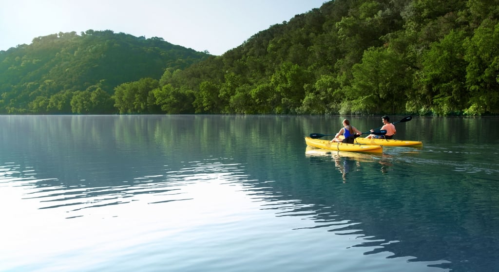 Two people kayaking on the water. In the background are two mountains flush with trees.