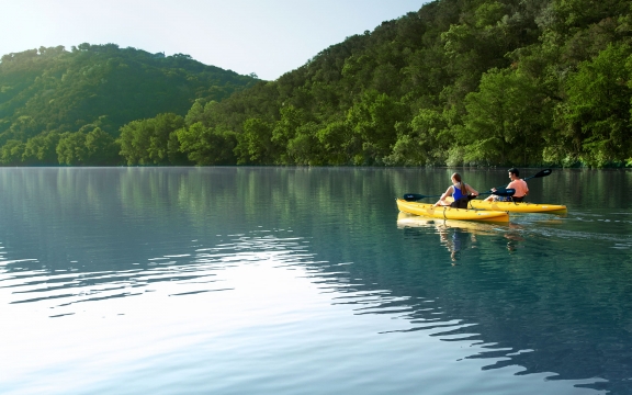 Two people kayaking on the water. In the background are two mountains flush with trees.