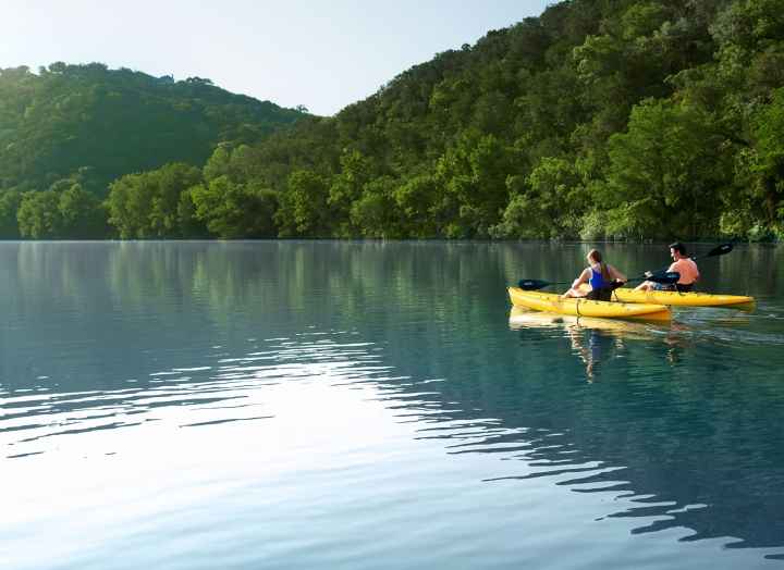 Two people kayaking on the water. In the background are two mountains flush with trees.