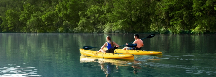 Two people kayaking on the water. In the background are two mountains flush with trees.