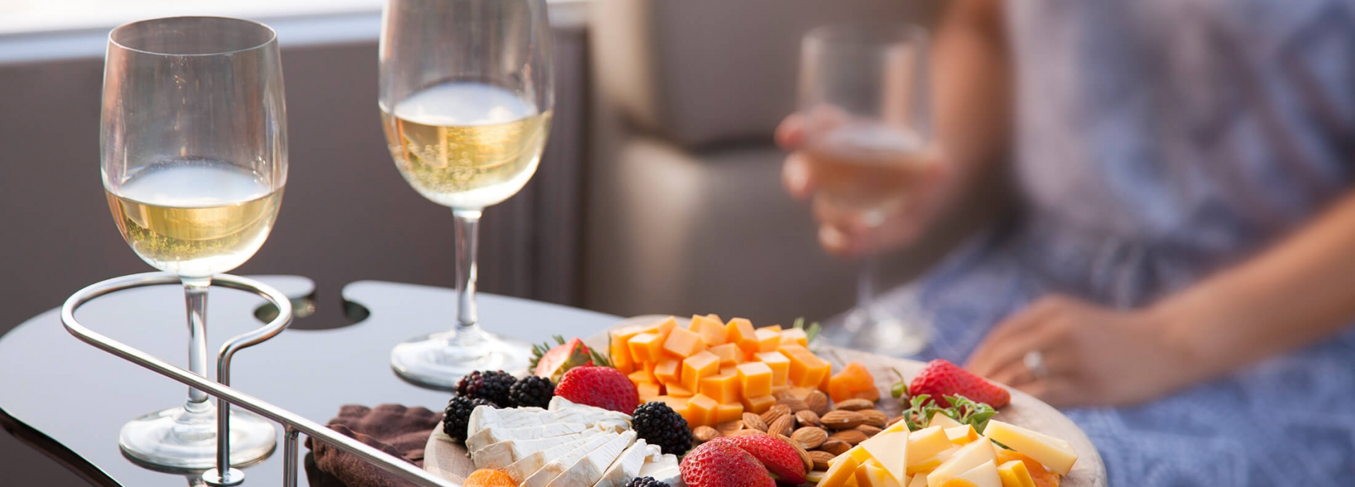 A woman sits at a tray of food consisting of cheeses, almonds, and berries alongside two glasses of wine.