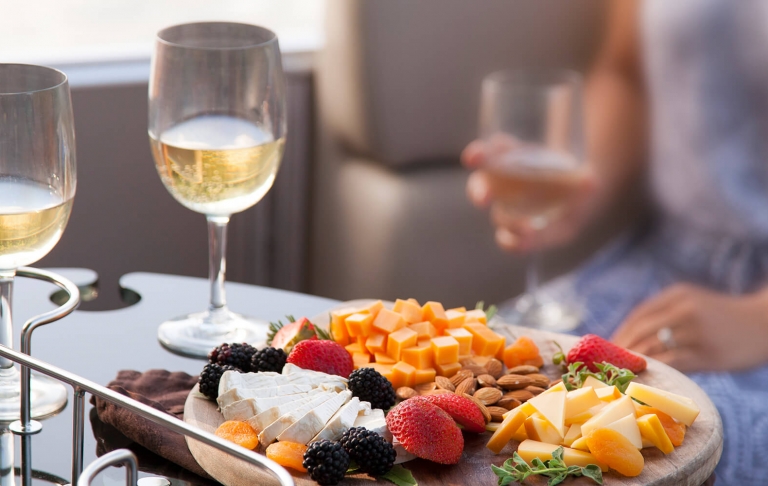 A woman sits at a tray of food consisting of cheeses, almonds, and berries alongside two glasses of wine.