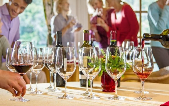 A group of guests socialize near a table where red wine is being poured into several glasses.
