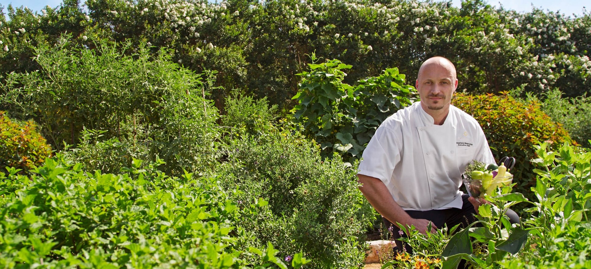 A chef poses for a photo in a beautiful green garden with a bowl of fresh vegetables in his hand.