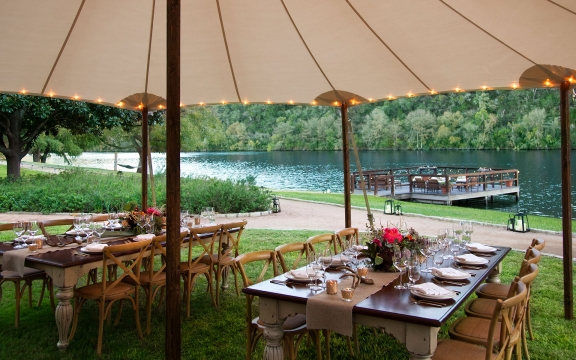 Two tables set for dinner under a tan umbrella. In the distance, additional tables can be seen on a dock by the water.