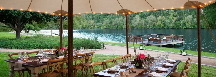 Two tables set for dinner under a tan umbrella. In the distance, additional tables can be seen on a dock by the water.