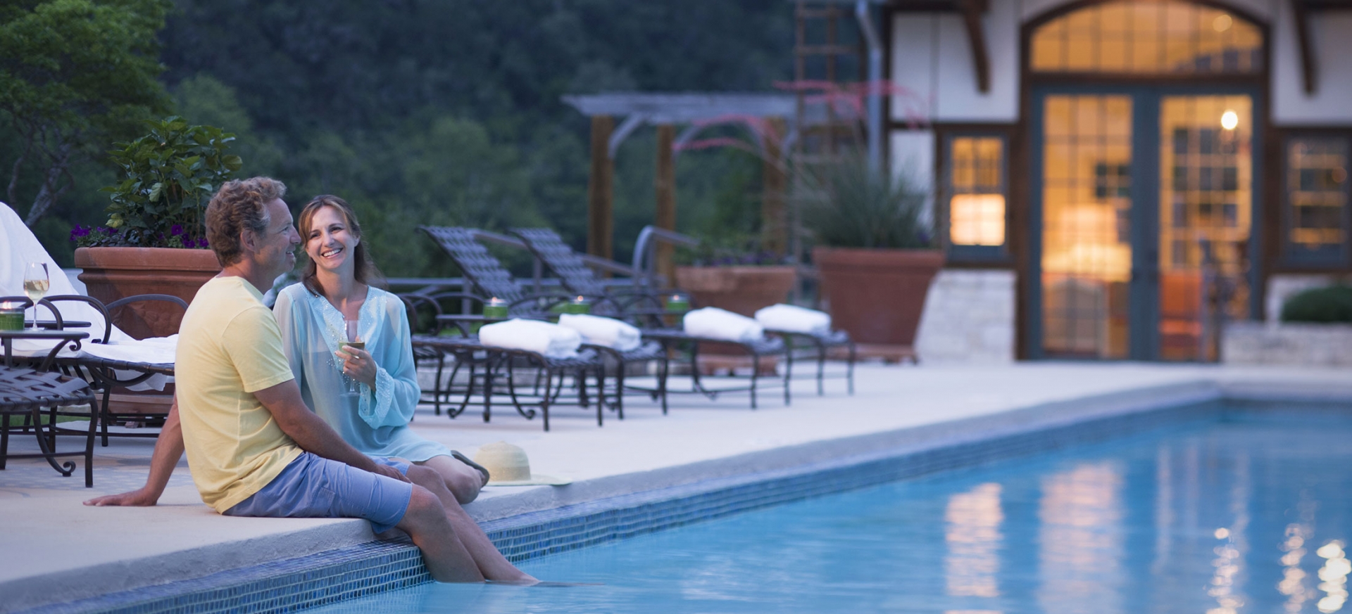 A couple sits poolside in the evening. The man smiles with his feet in the water. The woman smiles and holds a glass of wine.
