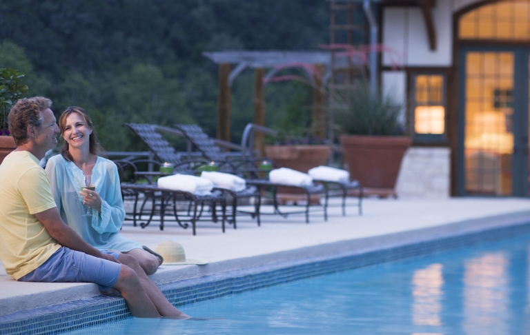 A couple sits poolside in the evening. The man smiles with his feet in the water. The woman smiles and holds a glass of wine.
