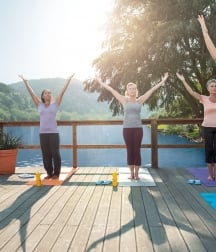 Three women attend a yoga class as their instructor guides them through a series of moves on a deck over the water.