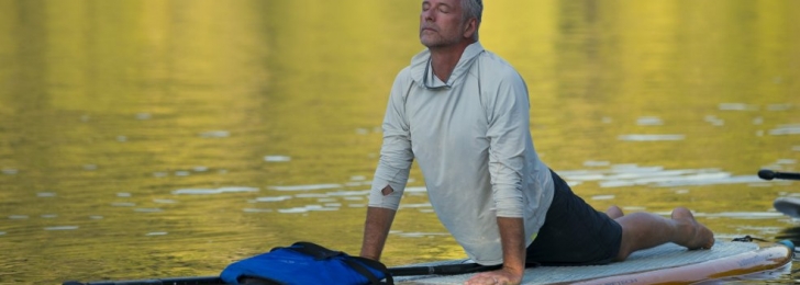 A man does yoga on a standup paddleboard on a calm lake. His body is face down but his torso stretches up into cobra pose.