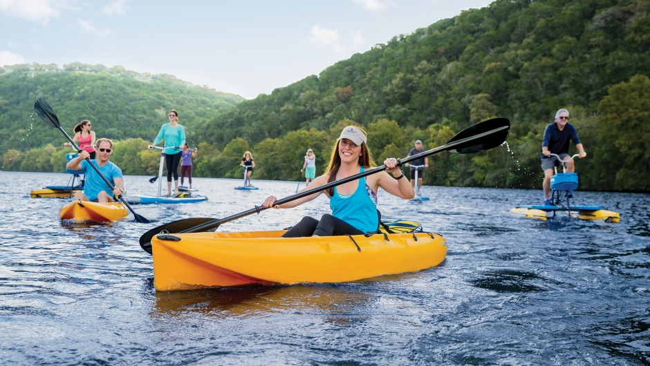 A group of people smiling on the lake enjoying paddling in kayaks, on standup paddleboards, pedal boards, and water sports