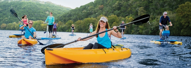 A group of people smiling on the lake enjoying paddling in kayaks, on standup paddleboards, pedal boards, and water sports