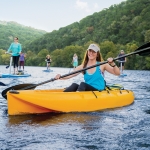 A group of people smiling on the lake enjoying paddling in kayaks, on standup paddleboards, pedal boards, and water sports