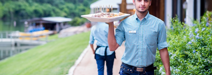 A male server in jeans and a nice checkered shirt carries a tray loaded with hors d'hoeuvres along an outdoor path.