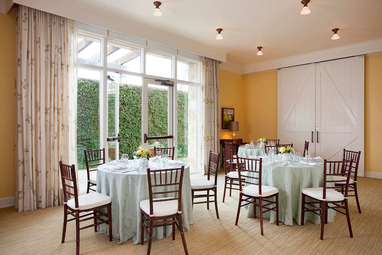 A brightly lit dining room with round tables set for a meal, large windows, and a view of a garden outside.