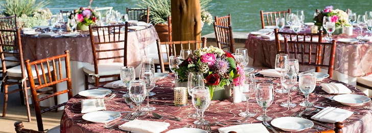 Three outoor dining tables fully set with glassware and utensils and wooden chairs. In the background a lake is visible.