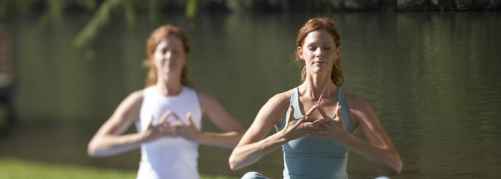 Two women sit together in meditation on the grass with a lake in the background.