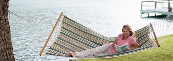 A woman laying in a striped green hammock overlooking the lake while reading a book.
