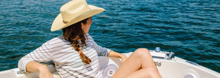 A woman in a striped shirt and cowboy hat sits at the front of a speedboat overlooking the water as the sun shines.