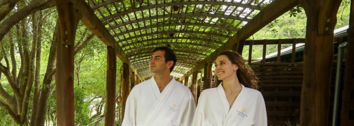 A couple walks under a wooded canopy surrounded by greenery in white Lake Austin branded robes