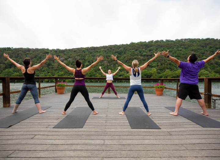 Four yoga students with black yoga mats stand facing their yoga teacher on a wharf overlooking the lake.