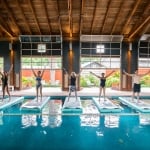 Four people stand on floating yoga mats in a large indoor pool with a view of green gardens in the background.