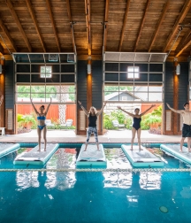Four people stand on floating yoga mats in a large indoor pool with a view of green gardens in the background.
