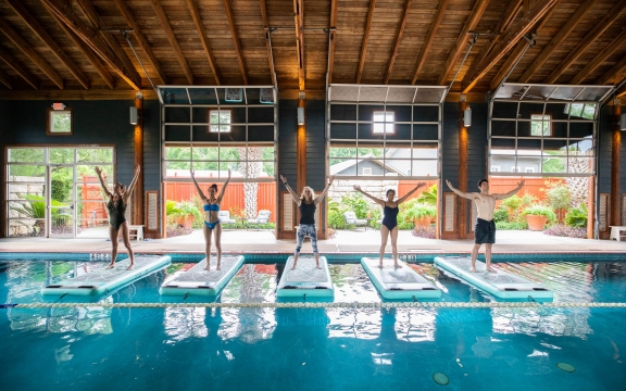 Four people stand on floating yoga mats in a large indoor pool with a view of green gardens in the background.