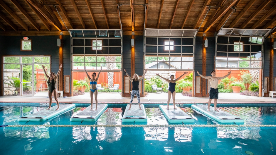 Four people stand on floating yoga mats in a large indoor pool with a view of green gardens in the background.
