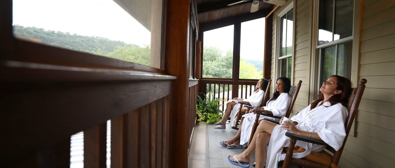 Three women sit on an outdoor porch in wooden rocking chairs all wearing white robes and blue sandals. One woman holds a mug.