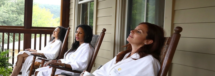 Three women sit on an outdoor porch in wooden rocking chairs all wearing white robes and blue sandals. One woman holds a mug.