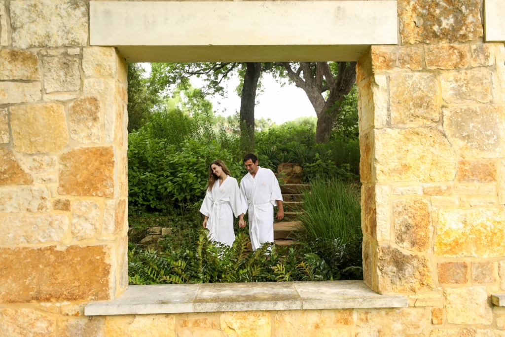 A tan stoned wall with a window where a couple can be seen wearing white robes while holding hands surrounded by greenery.