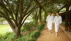 A couple walks under a wooded canopy surrounded by greenery in white Lake Austin branded robes