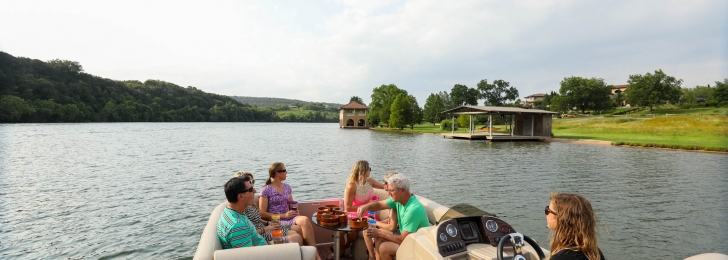 A group sits at the front of a boat sharing a meal while a woman drives. A gazebo and green hillsides can be seen from lake.