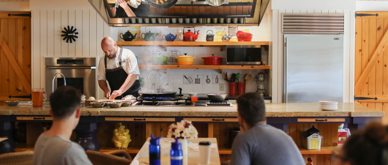 A chef in a black apron prepares food at a large island with colorful pots and pans in the background.