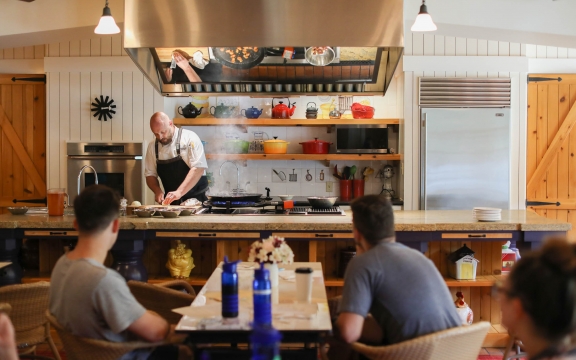 A chef in a black apron prepares food at a large island with colorful pots and pans in the background.