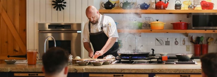 A chef in a black apron prepares food at a large island with colorful pots and pans in the background.