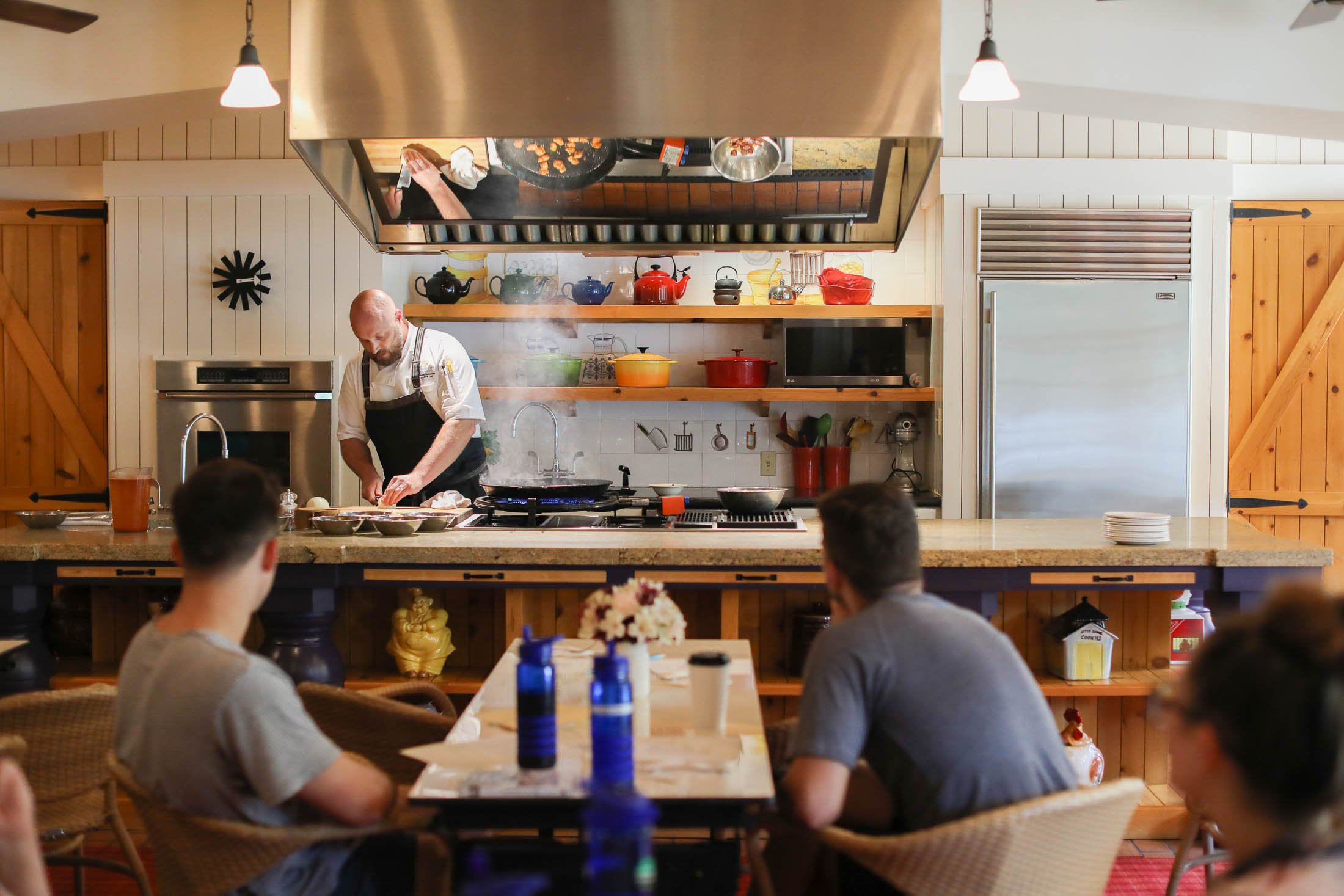 A chef in a black apron prepares food at a large island with colorful pots and pans in the background.