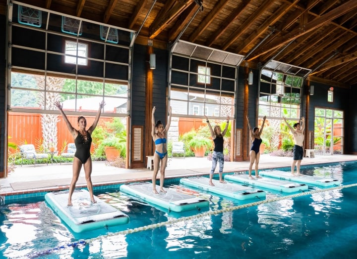 Five people stand on floating yoga mats in the indoor pool during a yoga class.