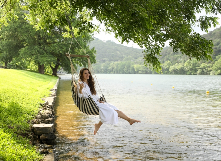 A woman sits in a hammock above the a large lake with a small grassy hill in the background.