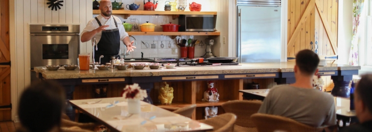 A chef stands at the front of the room with stainless steel kitchen appliances behind him teaching a class of students.