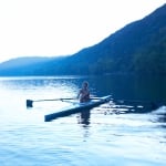 A woman sits in a single person rowboat on a lake at dawn.