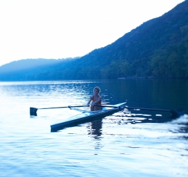 A woman sits in a single person rowboat on a lake at dawn.