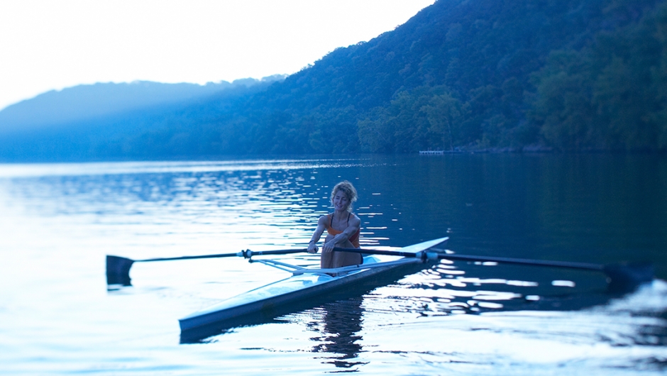 A woman sits in a single person rowboat on a lake at dawn.