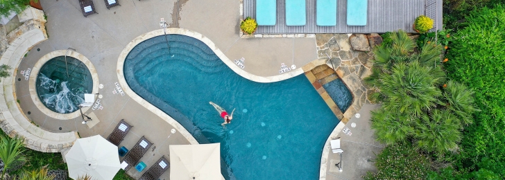 An aerial view of the pool with a woman floating inside it, a hot tub and pool chairs.