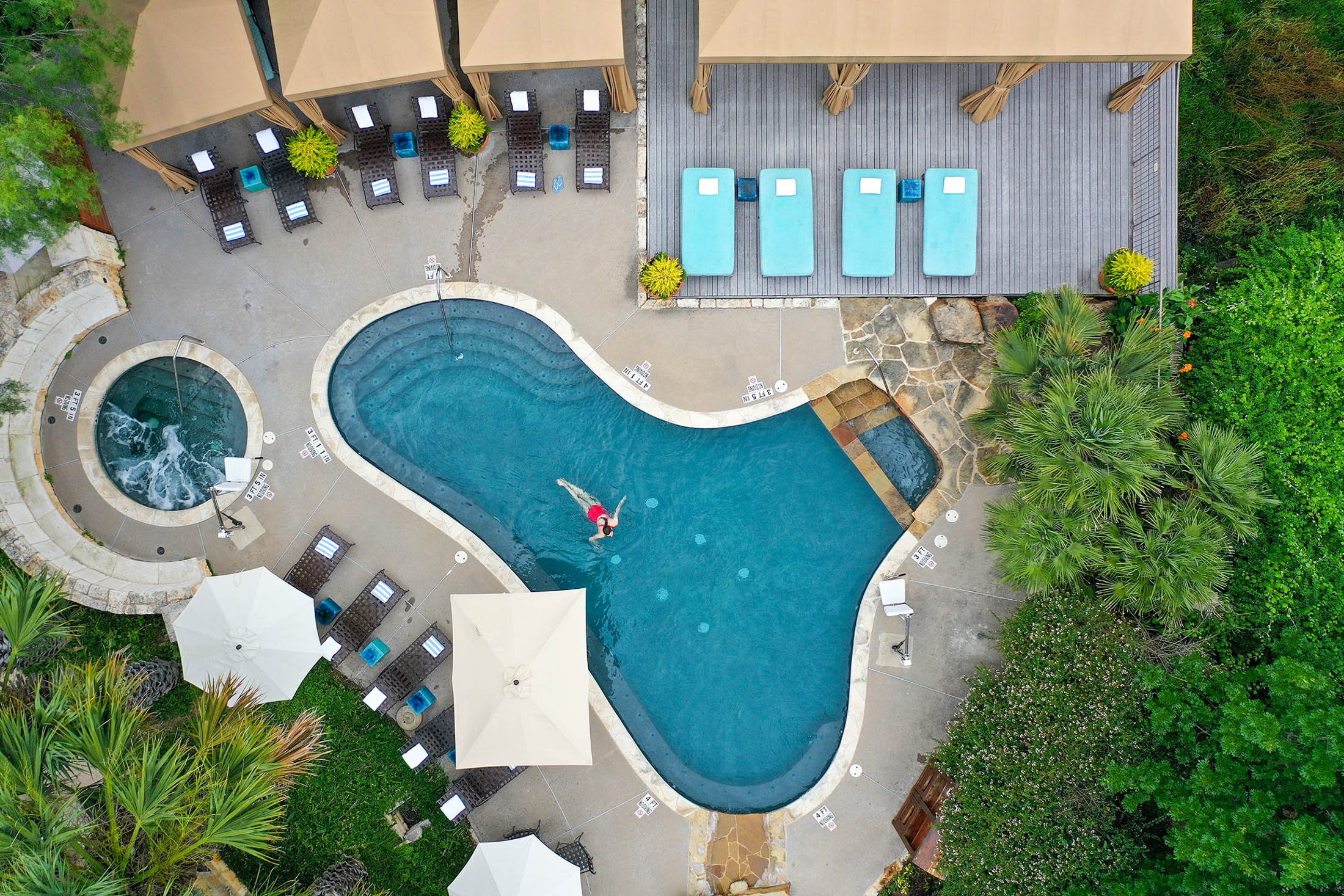 An aerial view of the pool with a woman floating inside it, a hot tub and pool chairs.