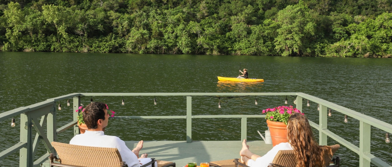 A couple sits on patio furniture on a green wharf facing the lake where a yellow sea kayak can be seen.