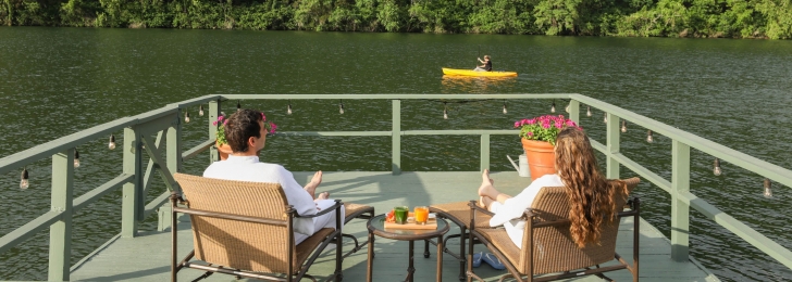 A couple sits on patio furniture on a green wharf facing the lake where a yellow sea kayak can be seen.