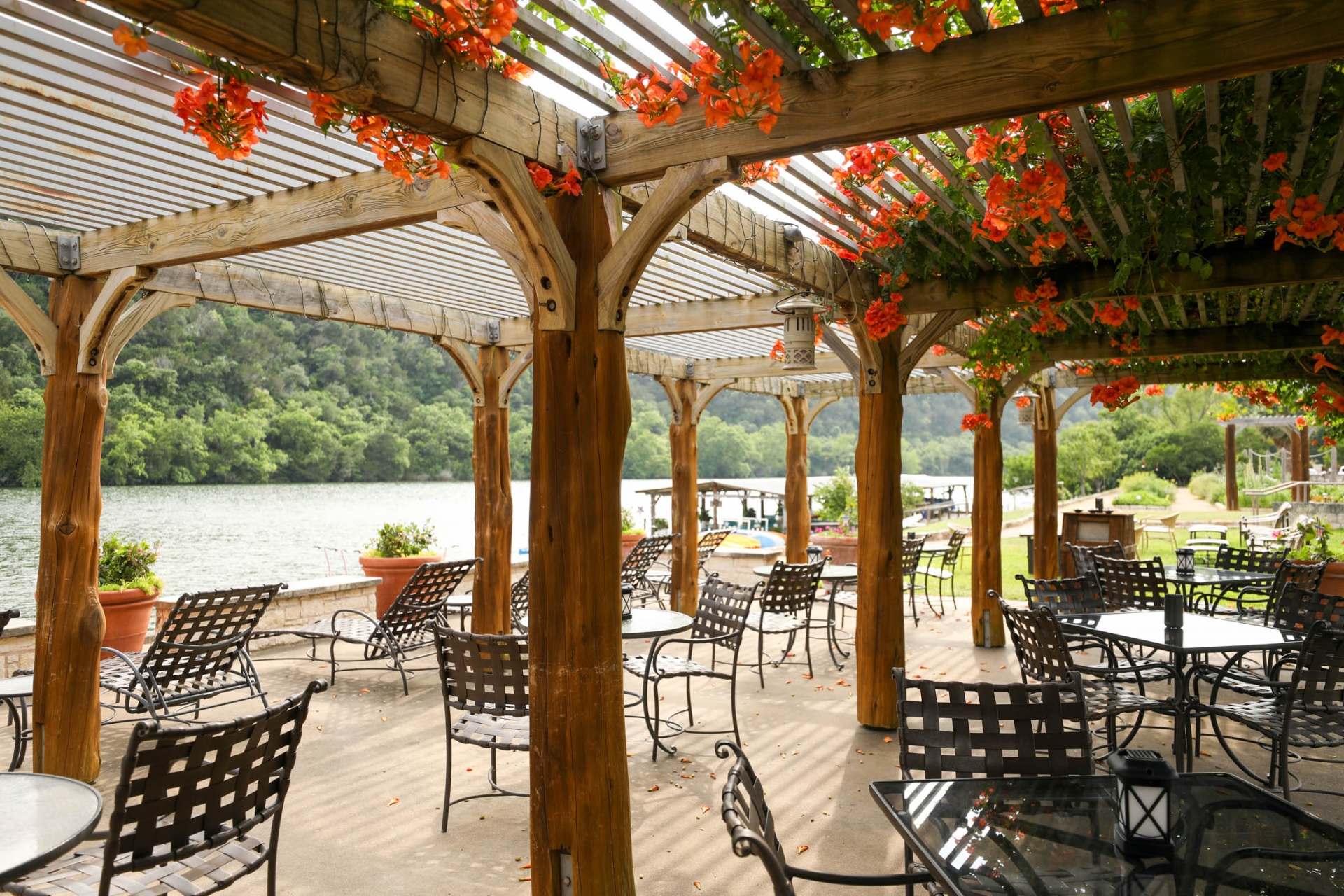 An outdoor patio where tables and chairs sit with a natural wood canopy and orange flowers peaking through its roof.